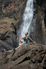 long hair man reclining on a large rock under a huge waterfall in Australia