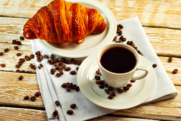 Coffee in a white ceramic mug. Croissant with coffee for breakfast. Coffee on an old wooden table. Coffee beans are scattered near the mug.