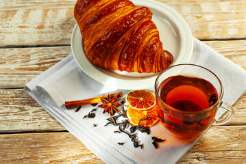 Croissant in a white plate with tea. Mug with tea on an old wooden table. Whole-leaf black tea, lemon and cinnamon on a napkin. Copy space and free space for text near food.