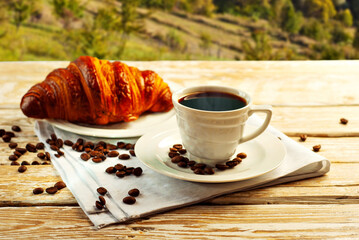 Coffee in a white ceramic mug. Croissant with coffee for breakfast. Coffee on an old wooden table. Coffee beans are scattered near the mug.