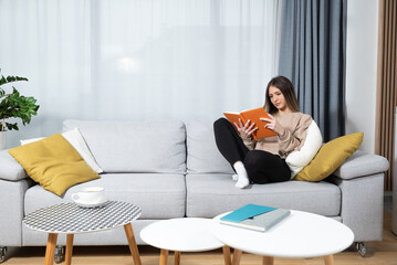Young beautiful woman sitting on the sofa at cozy home reading the book and enjoying her free time without her husband and children with smile and happy face. Female student study and relaxing.