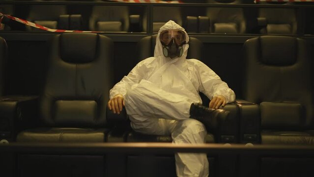 Relaxed man in white protection costume and respirator sitting alone at the cinema