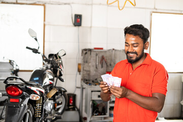 happy smiling mechanic couting money or current at garage - concept of successful small business, earning, financial and banking.