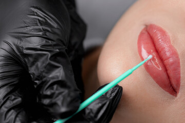 Master applies protect gel on red of lips young woman after permanent makeup tattoo in beautician...