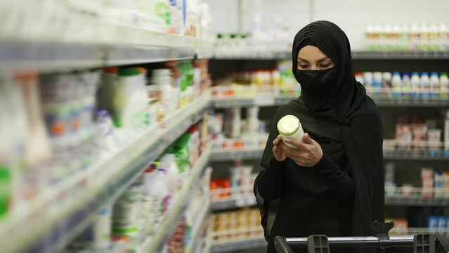 Woman in hijab and protective mask doing shopping, takes product from the shelf in a milk section