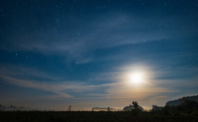 A beautiful summer night starry sky and the moon illuminates a misty field and forest, a nighttime natural landscape