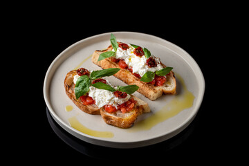 Two grilled bruschetta with stracciatella cheese and sun-dried tomatoes on black plate stand, top...