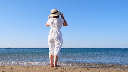 Fototapeta na wymiar back view on a mature woman in a straw hat and white dress walks along the blue sea coast on a sunny summer day, enjoying freedom and relaxation. The concept of a typical life of pensioners.