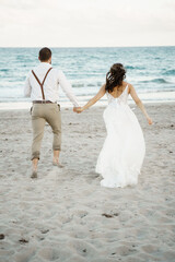Fototapeta na wymiar Bride in a white gown and groom in a white shirt running on the beach holding hands on the sandy shore Sea shore background
