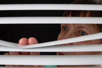 An elderly woman peers through the blinds through the window.