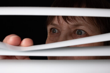 An elderly woman peers through the blinds through the window.