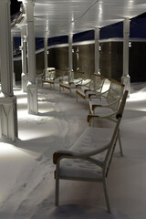 Winter. Evening. The benches standing in a semicircle are covered with snow. Beautifully illuminated architectural canopy with columns. 