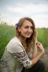 Fototapeta premium Girl wearing national Ukrainian clothes embroidered shirt is sitting in the beautiful rye field. Woman is holding blue field flowers cornflowers. Blue wildflowers and blond wavy haired girl 
