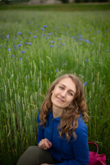 Satisfied blond girl is sitting on green grass near blue wildflowers. Women is wearing dark blue hoodie near cornflowers on the green background.
