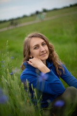 Satisfied blond girl is sitting on green grass near blue wildflowers. Women is wearing dark blue hoodie near cornflowers on the green background.
