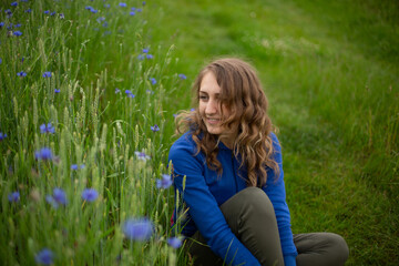Fototapeta premium Satisfied blond girl is sitting on green grass near blue wildflowers. Women is wearing dark blue hoodie near cornflowers on the green background. 