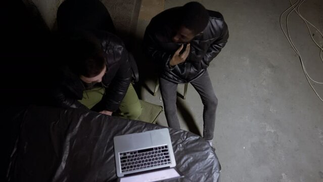 Caucasian guy and black guy are sitting near laptop in a basement during a bombardment and having a talk, russian invasion in Ukraine