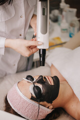 On the skin of the girl's face, gel with carbon was applied and black goggles were worn. The cosmetologist holds a laser in his hands and performs a carbon peeling procedure. 