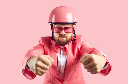 Funny Furious Tough Driver Holding Pretend Steering Wheel. Studio Shot Of Man With Ginger Beard And Angry Face, Wearing Pink Helmet, Funky Modern Suit, Bow Tie And Sunglasses Driving His Invisible Car