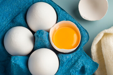Close-up of chicken fresh eggs in eco-packaging on a blue background. Broken egg with yolk in the shell. Farm natural products. Top view. - obrazy, fototapety, plakaty