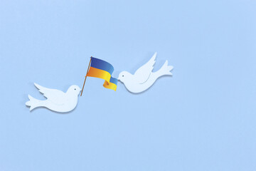 Two white figurines of a doves of peace with  flag of Ukraine  in their beaks on the light blue background. Peace Concept