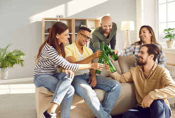 Smiling multiracial young people clink bottles relax at home together watching TV or football game....