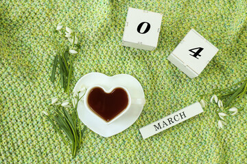 Calendar for March 4: cubes with the numbers 0 and 4, the name March in English, a cup of tea in the shape of a heart, snowdrops on a knitted knitted fabric, top view