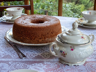 Brown cake on a table with white KPM porcelain tea set, decorared with flowers. White lace tablecloth, silver knife, sugar pot. Behind, a window with view to the garden.