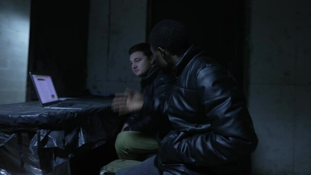 Caucasian guy and black guy are sitting in a basement and discussing war news from a laptop