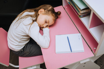 A beautiful, little red-haired girl, a child, a schoolgirl sits at a wooden desk at school and sleeps, in a lesson. Photography, concept.