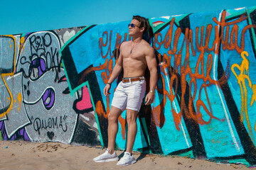 Slim young muscular male model with no shirt and white shorts, on a summer day at the beach next to a blue graffiti wall in Venice California.
