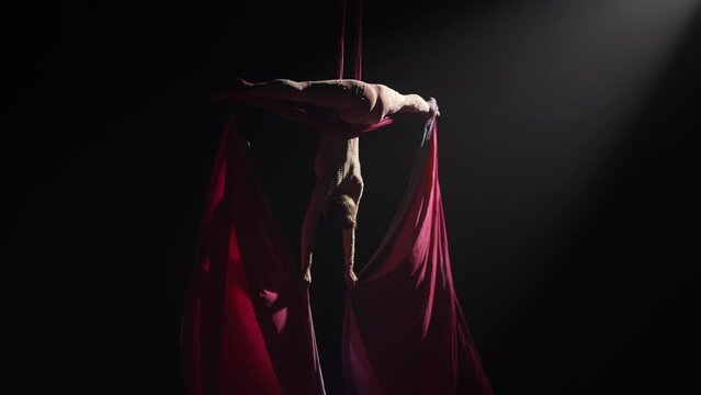 Young female circus gymnast spins hanging upside down on aerial silk. Acrobat performs tricks at height, red silk flutters like wings on black background with backlight. Slow motion.