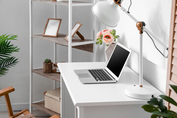 Modern workplace with laptop, flowers in vase and lamp near light wall