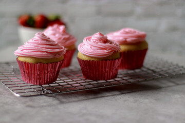 vanilla muffins decorated with pink buttercream frosting on top in red baking cups, on grey marble table, strawberries and white stone wall in background 