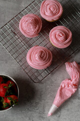 view from above on muffins with pink buttercream frosting on top on grey marble table with strawberries and pastry bag 