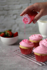 hand strews pink sprinkles on muffins with pink buttercream frosting on top in red baking cups, on grey marble table, strawberries and white stone wall in background 