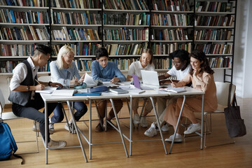 Full length concentrated team of multiethnic millennial college university students studying sitting together at big table, working on project in library using gadgets and paper books, writing notes.