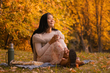 Cozy young Mongolian asian woman smiling with her eyes closed enjoying a wonderful cup of tea on picnic in a park in autumn. 