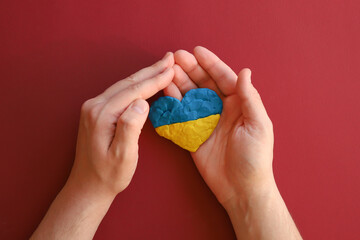 Ukrainian Ukraine flag, no war. Heart shape of blue and yellow plasticine modeling clay in male hands on red maroon texture background. Plasticine finger textured background. Top view, closeup.