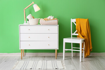 Fototapeta premium Chest of drawers with baby clothes and chair with knitted sweater near color wall in room interior