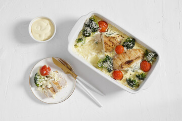 Baked chicken fillet with broccoli and tomatoes in a white form, served with portion on the plate and cutlery on a white background, keto diet, top view
