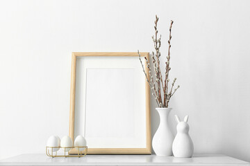Blank photo frame, holder with Easter eggs and pussy willow branches on table near light wall