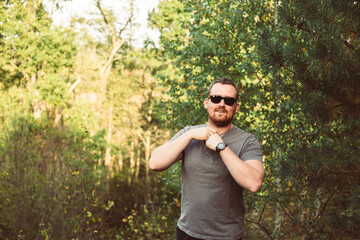 Young strong man in gray t-shirt and black sunglasses posing outdoors in the green summer forest