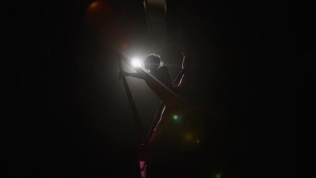 Acrobatic performance of a young woman circus performer on airy silk. Equilibrium circus gymnast spinning in the air on a black backlit background, low angle. Slow motion.