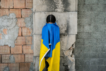 Little girl with Ukrainian flag in front of a wall destroyed from bombs. The little girl waves the...