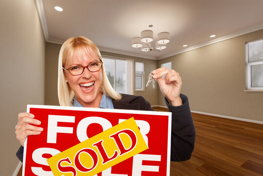 Attractive Young Woman with New Keys and Sold Real Estate Sign In Empty Room of House