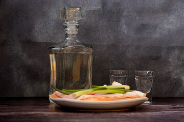 A bottle of vodka, two glasses and a plate with two sandwiches with bacon and green onions on a wooden table.