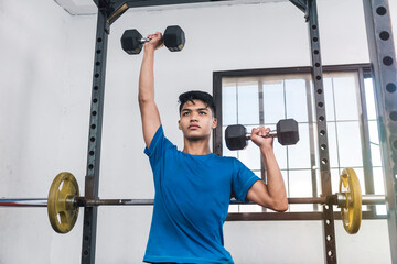 A young asian man wearing a blue shirt and seated on a bench does alternating dumbbell presses....