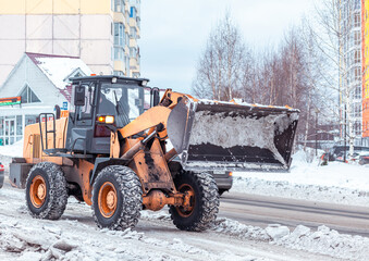 Big orange tractor cleans up snow from the road and loads it into the truck. Cleaning and cleaning of roads in the city from snow in winter. Snow removal after snowfall and blizzards. 