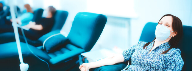 Woman in medical mask sits in chair and donates blood in medical clinic. Topic of donation. Donor...